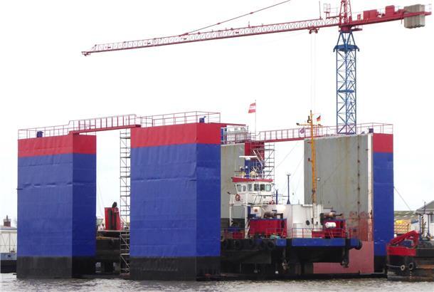 Heise-Werft nimmt 34 Meter langes Schwimmdock „Wilhelmina“ in Betrieb Ein schwimmendes Trockendock mit blau-roter Bemalung im Wasser, vor einem Kran im Hintergrund.