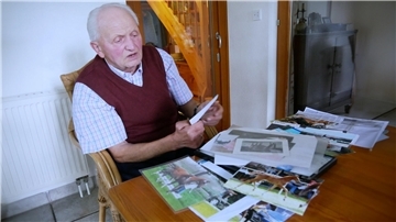 Ein alter Mann sitzt an einem Tisch, vor ihm ist ein Stapel mit Fotos.