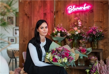 Eine Frau, die eine schwarze Hose und Weste über einem weißem Pullover trägt. Sie hält einen Blumenstrauß in der Hand. Hinter ihr sind noch weitere Blumensträuße zu sehen.