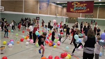 Sportfest in einer Turnhalle mit vielen Kindern und Luftballons.