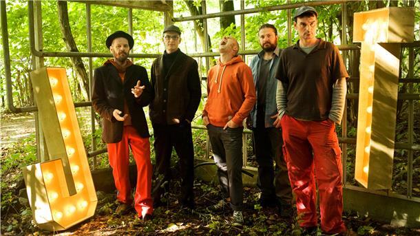Eine fünfköpfige Band steht in einem bewaldeten, grünen Außenbereich vor einer Metallkonstruktion. Die Mitglieder tragen legere Kleidung, einige mit roten Hosen, Hüten oder Kapuzenpullovern. Links und rechts im Bild leuchten große, beleuchtete Buchstaben „J“ und „T“.