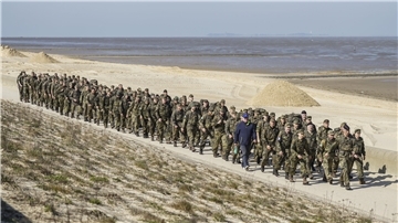 Eine große Gruppe von Soldaten in Tarnuniform marschiert in Reih und Glied an einem Deich entlang, im Hintergrund ist die Nordseeküste mit Sandflächen und Meer zu sehen