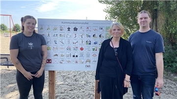 Drei Personen stehen am Strand vor einer Tafel mit Piktogrammen.