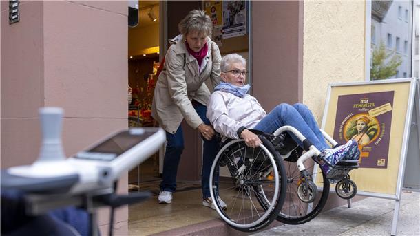 Eine Frau hilft einer Rollstuhlfahrerin beim Weg in ein Café.