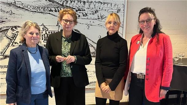 Vier Frauen stehen nebeneinander in einem Innenraum vor einer großformatigen historischen Zeichnung. Sie tragen unterschiedliche Business- und Freizeitkleidung, darunter Blazer, Blusen und ein rotes Jackett.