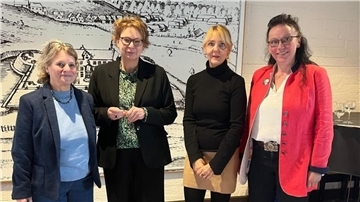Vier Frauen stehen nebeneinander in einem Innenraum vor einer großformatigen historischen Zeichnung. Sie tragen unterschiedliche Business- und Freizeitkleidung, darunter Blazer, Blusen und ein rotes Jackett.