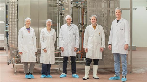 Zwei Frauen und drei Männer mit Hauben und weißen Kitteln stehen vor einer industriellen Anlage.