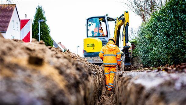 Arbeiter sind mit Bagger und Spaten an einer Straße mit der Erdverlegung von Glasfaserkabeln beschäftigt.