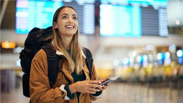 Junge Frau steht im Flughafen und trägt einen schwarzen Rucksack auf dem Rücken und hält ein Smartphone in der Hand. 