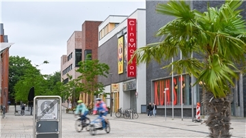 Blick auf das CineMotion-Kino in Bremerhaven mit rotem Fassadenschild. Im Vordergrund eine Palme und Radfahrende in Bewegung, rechts das Kunstmuseum mit großer „KUNST“-Beschriftung im Fenster.
