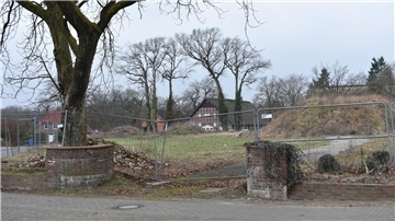 Auf einer Brachfläche mit einem Bauzaun  sind vorne die Reste einer Backsteinmauer und ein hoher Baum zu sehen.