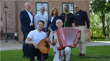 Sieben Personen posieren vor der Backsteinfassade der Burg Bederkesa. Zwei Musiker knien vorn mit Mandoline und Akkordeon, fünf weitere Personen stehen dahinter auf dem Rasen.