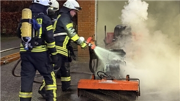 Drei Männer in schwarz- und signalgelben Anzügen halten einen Wasserstrahl auf einen brennenden Aufsitzrasenmäher.