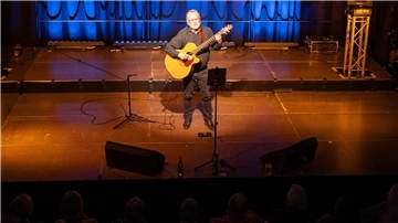 Ein Mann steht mit Gitarre auf einer stimmungsvoll beleuchteten Kleinbühne.