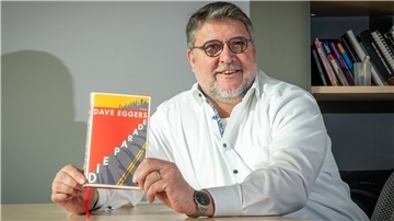 Ein Mann sitzt an einem Tisch und zeigt das Buch "Die Parade" in die Kamera.