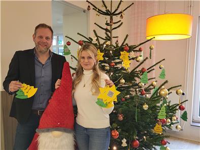 Ein Mann und eine Frau stehen mit Wunsch-Tannenbäumen in der Hand vor einem geschmückten Tannenbaum mit weiteren Wünschen. 