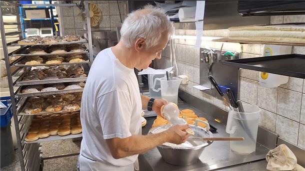Wie Bäcker in Elsdorf den Tag beginnen – Ein Blick in die Backstube Schwarz Ein Mann glasiert Berliner in einer Backstube. Im Hintergrund sind viele Kuchen auf Blechen.