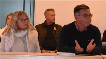 Eine Frau und ein Mann sitzen in einem Innenraum an einem Tisch. Die Frau trägt eine Brille, einen hellen Schal und eine Jacke. Der Mann neben ihr trägt ebenfalls eine Brille, einen dunklen Pullover über einem Hemd. Er gestikuliert mit seinen Händen. Im Hintergrund sitzen weitere Personen.