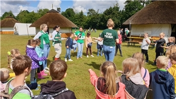 Kinder auf einer Wiese, die einen Kreis um eine erwachsene Frau gebildet haben.
