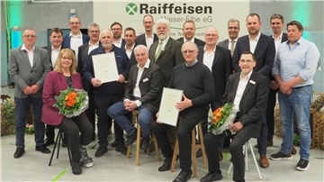 Eine Gruppe von Männern und Frauen posiert in einer Veranstaltungshalle vor einem Banner mit der Aufschrift „Raiffeisen Weser-Elbe eG“. Einige Personen in der ersten Reihe halten gerahmte Ehrenurkunden und Blumensträuße. 