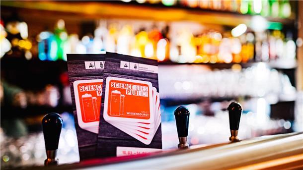 Bremerhaven feiert 20 Jahre „Stadt der Wissenschaft“ Zwei Flyer der Veranstaltungsreihe „Science goes Public!“ stehen auf einer Theke in einer Bar, im Hintergrund unscharf bunte Flaschen und Zapfhähne.