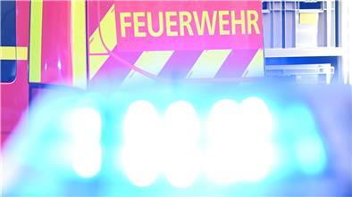 Ein Fahrzeug der Feuerwehr steht bei einem Einsatz auf einer Straße, während im Vordergrund das Blaulicht eines Polizeifahrzeugs zu sehen ist. 