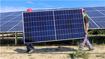 Zwei Männer tragen ein Solarpanel. Im Hintergrund ist ein Solarpark zu sehen.