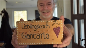 Ein Mann hält ein Schild in die Kamera. Darauf steht "Lieblingskoch Giancarlo" geschrieben.