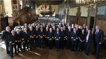 Gruppenfoto von Kapitänen und Gästen in Uniform und festlicher Kleidung beim 58. Bremer Kapitänstag in der historischen Oberen Rathaushalle mit prunkvollem Holzinterieur und großer Schiffsreplik an der Decke. Im Hintergrund sind festlich gedeckte Tische zu sehen.
