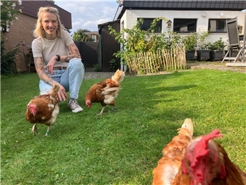 Eine blonde Frau hockt in ihrem Garten mit drei Hühnern.
