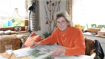 Eine Frau sitzt am Tisch und hält eine Zeitung in Händen.