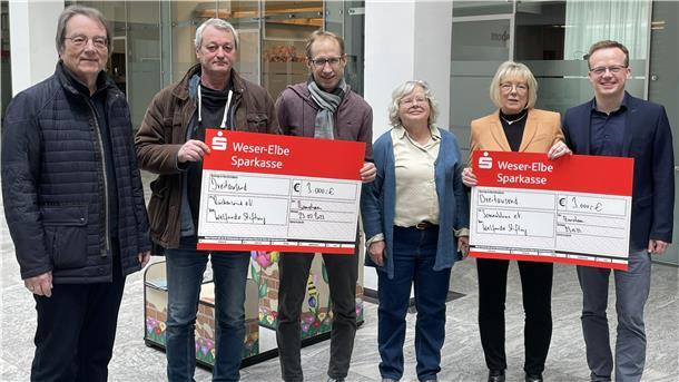 Eine Gruppe (sechs Menschen) steht vor einem Gebäude. Zu sehen sind auch zwei symbolische Schecks. 