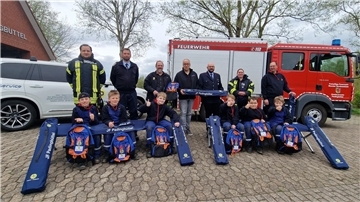 Vor einem Pkw und einem Einsatzfahrzeug der Feuerwehr Padingbüttel posieren sieben Erwachsene und sechs Kinder für ein Gruppenfoto mit neuen Liegen und Rucksäcken.