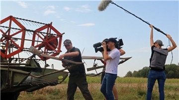Ein dreiköpfiges Filmteam mit Kamera und Mikrofon filmt einen Mann bei den Arbeiten an einem historischen Mähdrescher auf einem Feld.