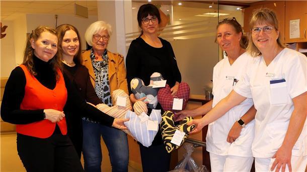 Sechs Frauen stehen in einem Krankenhausraum und halten bunte herzförmige Kissen in den Händen. Zwei der Frauen tragen Pflegekleidung, die anderen Alltagskleidung. Es werden Kissen übergeben.