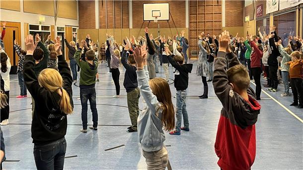 Körpertrommeln: Workshop für 1000 Grundschüler Eine große Gruppe von Kindern und Jugendlichen steht in einer Turnhalle und führt gemeinsam eine Übung aus. Alle haben die Arme nach oben gestreckt, während sie in Reihen aufgestellt sind.