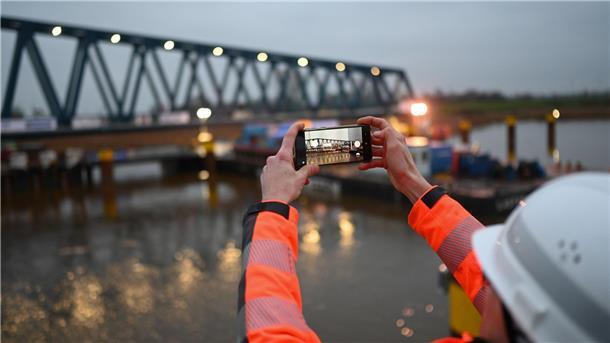 Jemand mit Helm und Arbeitsjacke fotografiert mit seinem Handy die neue Friesenbrücke bei Weener.