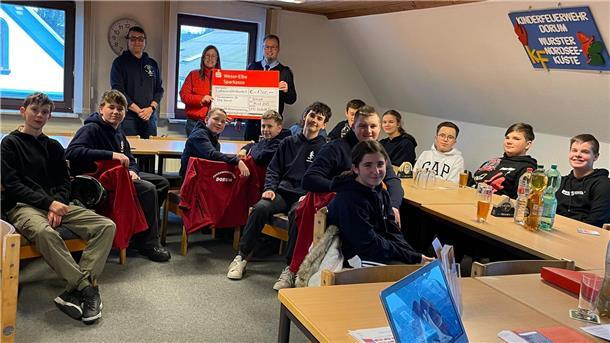 Jugendfeuerwehr Dorum erhält Spende aus Tannenbaumsammlung Eine Gruppe von Jugendlichen und jungen Erwachsenen sitzt in einem Gemeinschaftsraum an Tischen und Bänken. Viele tragen dunkle Hoodies mit Feuerwehr-Logos. Zwei Personen im Hintergrund halten einen großen symbolischen Spendencheck der Weser-Elbe Sparkasse über 1.300 Euro. An der Wand hängt ein Schild mit der Aufschrift „Kinderfeuerwehr Dorum Wurster Nordsee Küste“.