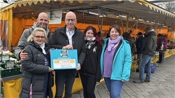 Fünf Personen stehen auf einem Wochenmarkt vor einem Gemüsestand und halten gemeinsam einen großen Spendenscheck über 1.000 Euro in die Kamera. Im Hintergrund sind Marktstände mit gelben Planen und weitere Besucherinnen und Besucher zu sehen.