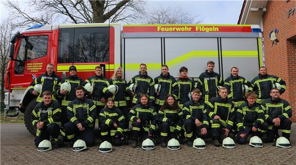 Gruppenfoto der Freiwilligen Feuerwehr vor einem Einsatzfahrzeug der Feuerwehr Flögeln. Die Feuerwehrleute tragen vollständige Schutzkleidung, viele halten ihre Helme in den Händen oder haben sie vor sich auf den Boden gestellt. Im Hintergrund sind das Feuerwehrfahrzeug und ein Teil des Feuerwehrhauses zu sehen.
