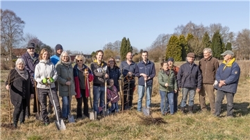 Eine Gruppe von 16 Menschen – von Kindern bis Senioren – steht auf einer Wiese an einem sonnigen Tag. Sie halten Schaufeln und junge Bäume.