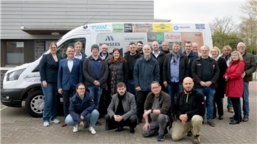 Gruppenfoto von rund 25 Personen vor einem weißen Transporter mit zahlreichen Sponsorenlogos; die Gruppe steht lächelnd auf einem Parkplatz vor einem modernen Gebäude.