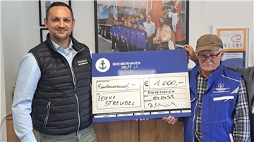 Zwei Männer stehen nebeneinander und halten gemeinsam einen überdimensionalen blauen Scheck über 1.000 Euro zugunsten von „Bremerhaven Hilft e.V.“, im Hintergrund hängt ein Bild von Jugendfußballern.