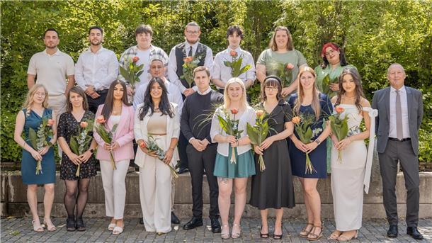 Gruppenfoto von 18 jungen Erwachsenen, die ihre Ausbildung zu Verwaltungsfachangestellten abgeschlossen haben. Sie stehen in zwei Reihen im Freien, halten Blumen in der Hand und lächeln in die Kamera. Rechts im Bild steht ein Mann im Anzug, vermutlich ein Vertreter des Magistrats. Im Hintergrund ist grünes Laub zu sehen, die Szene wirkt festlich und sommerlich.