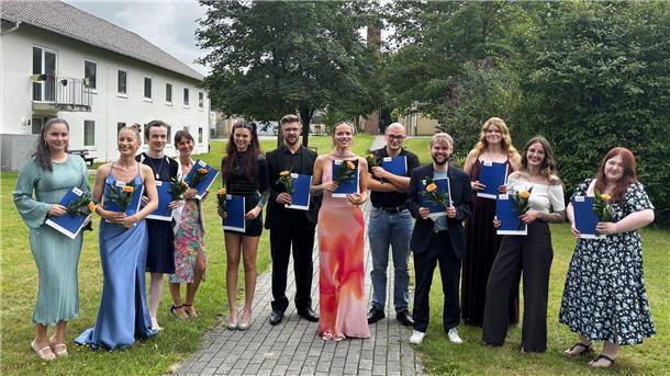 Zwölf neue Pflegefachkräfte feiern ihren Abschluss Zwölf frisch examinierte Pflegefachkräfte stehen mit Zeugnismappen und Blumen in der Hand lächelnd nebeneinander auf einem Weg im Grünen vor dem AMEOS Klinikum.