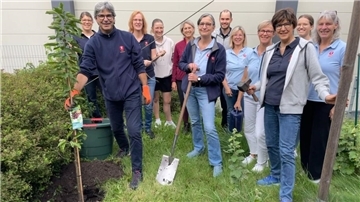 Eine Gruppe Männer und Frauen steht zusammen. Einige haben Werkzeug in der Hand und einer einen Baum.