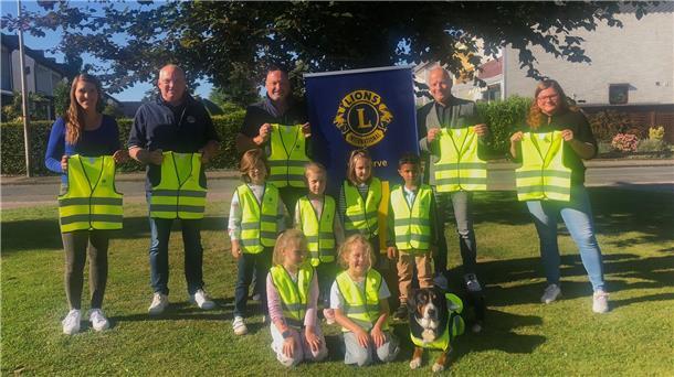 Lions Club spendet 250 Warnwesten für Erstklässler Mehrere Erwachsene und Kinder stehen und knien auf einer Wiese, alle tragen oder halten leuchtend gelbe Sicherheitswesten. Hinter ihnen ist ein Banner des Lions Clubs zu sehen. Auch ein Hund trägt eine Warnweste.
