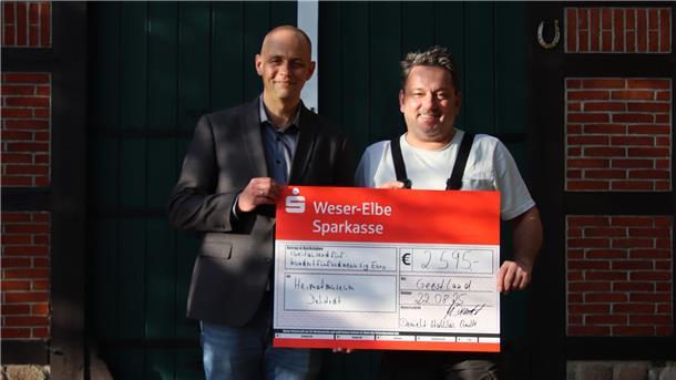 Zwei Männer stehen vor einem grünen Tor mit Fachwerkrahmen und halten gemeinsam einen großen symbolischen Spendenscheck der Weser-Elbe Sparkasse. Der Scheck ist auf das Heimatmuseum Debstedt ausgestellt und zeigt eine Spendensumme von 2.595 Euro.