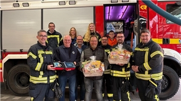Gruppenbild vor einem Einsatzfahrzeug der Feuerwehr Langen. Menschen halten einen Defibrillator und Präsentkörbe in den Händen.