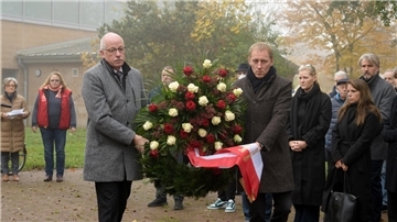 Zwei Männer tragen einen Kranz mit roten und weißen Blüten. Im Hintergrund stehen Menschen.